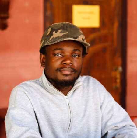 Headshot of Mohammed 'Ayo' Busari wearing a camo hat and a light grey jumper