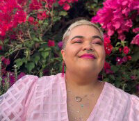 Smiling woman wearing pink stands against pink flowers and lush green background.