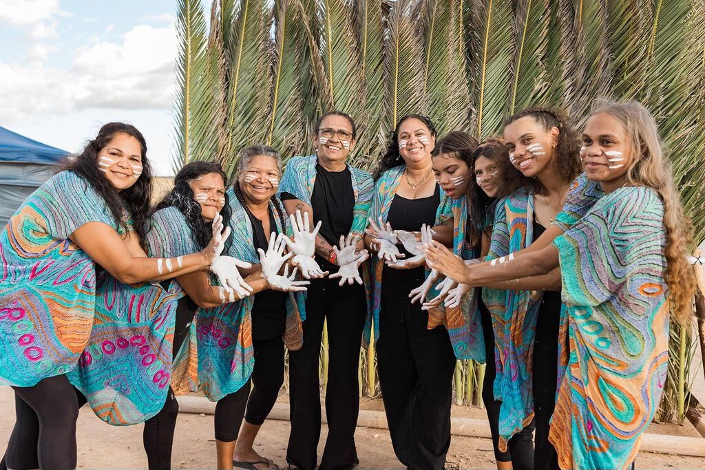 Group stands before tall grass wearing colorful patterned garments; white paint on hands and arms is prominently displayed.