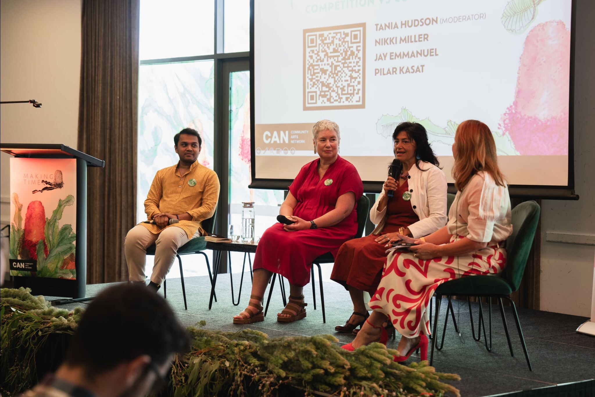 Four panelists sit on stage with a screen showing names and a QR code; podium and large windows in background.