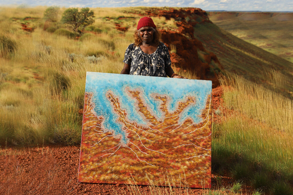 Person stands in grassy hills holding abstract painting with blue and brown branching patterns.