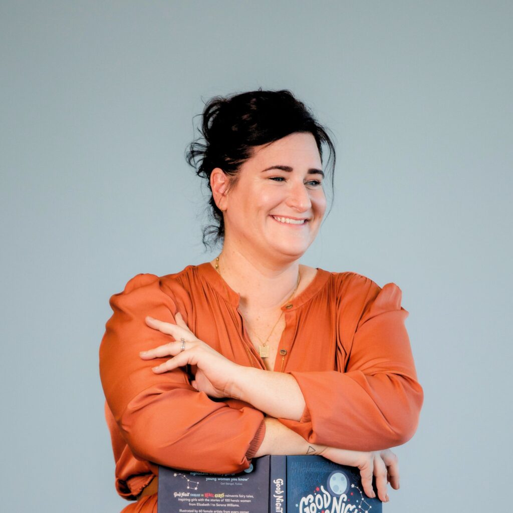 Woman in orange blouse holding the book "Good Night Stories for Rebel Girls" against plain background.