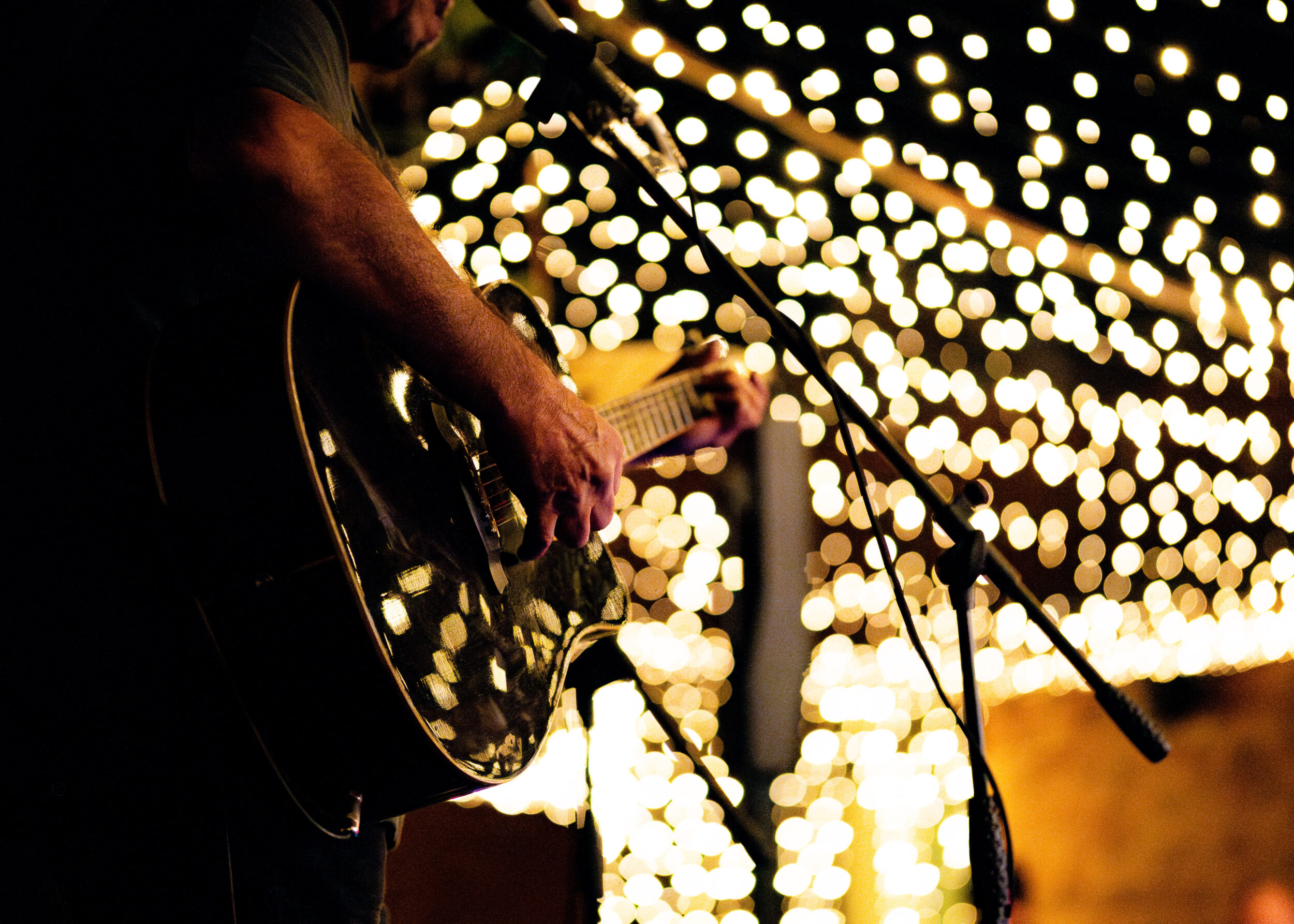 Close-up of a person playing an acoustic guitar on stage with a microphone in front of them. The background features out-of-focus string lights creating a bokeh effect.