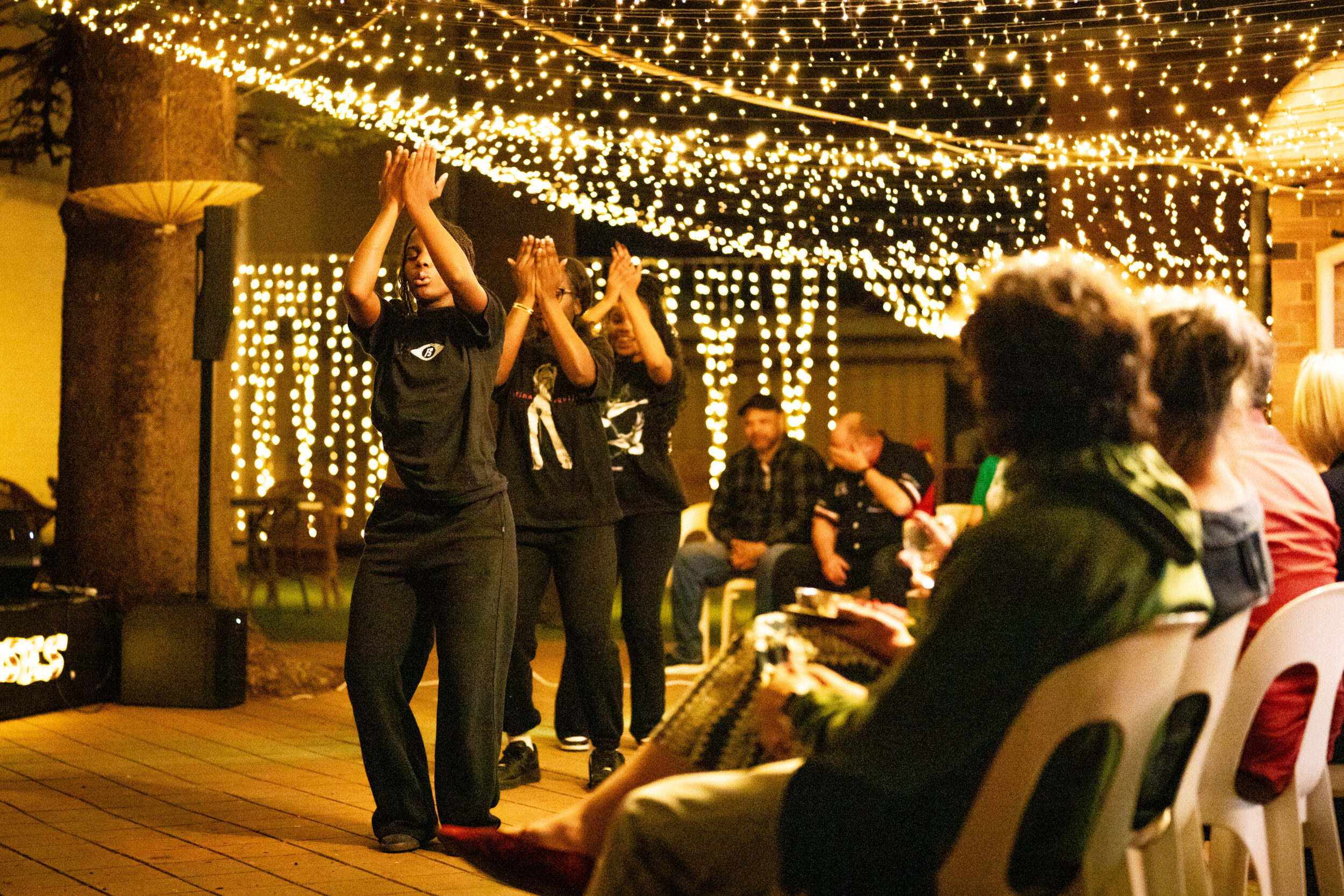 Audience watches three black-clad performers clapping on festive stage with string lights.