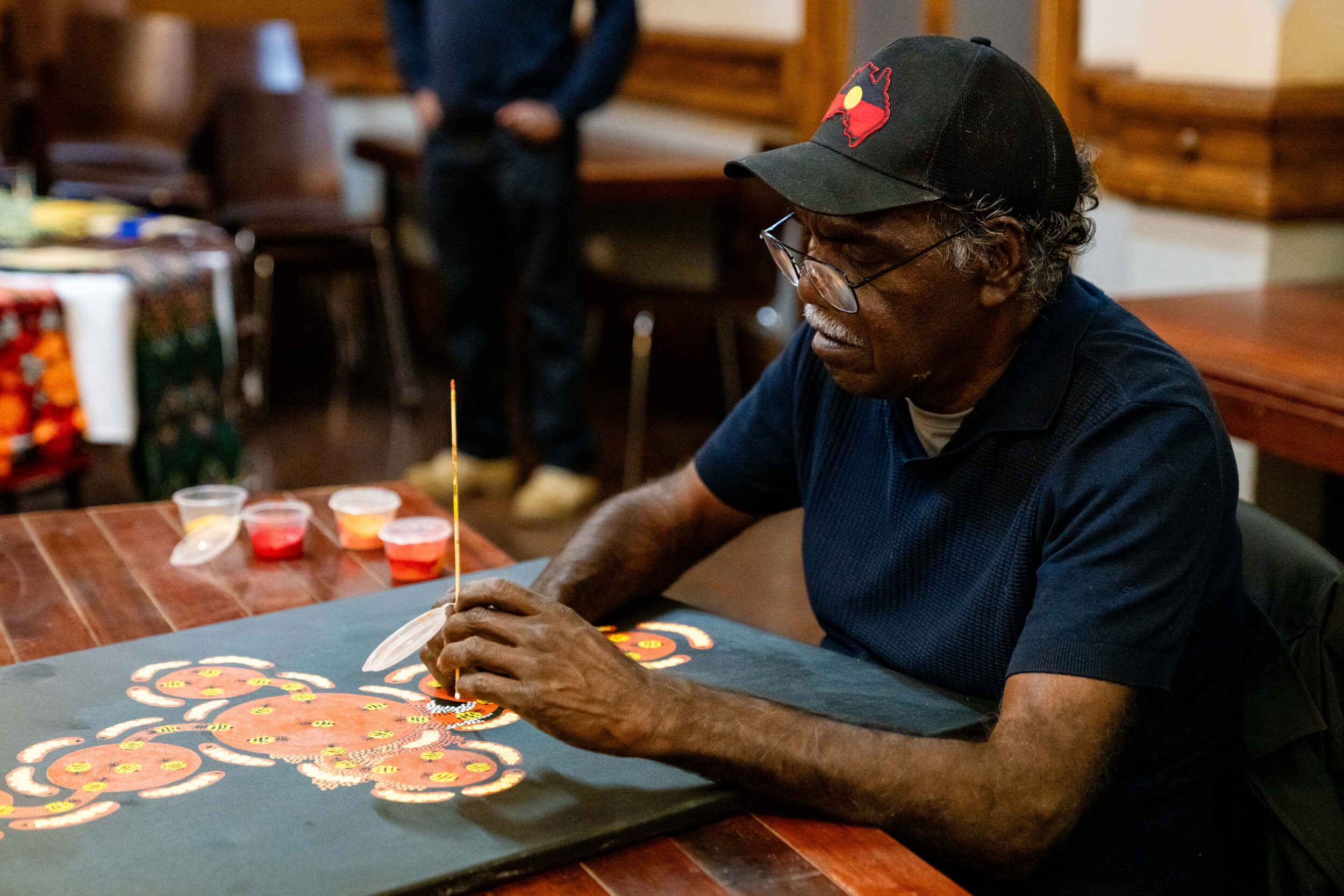 First Nations man wearing a hat is seated inside at a table doing a dot work artwork