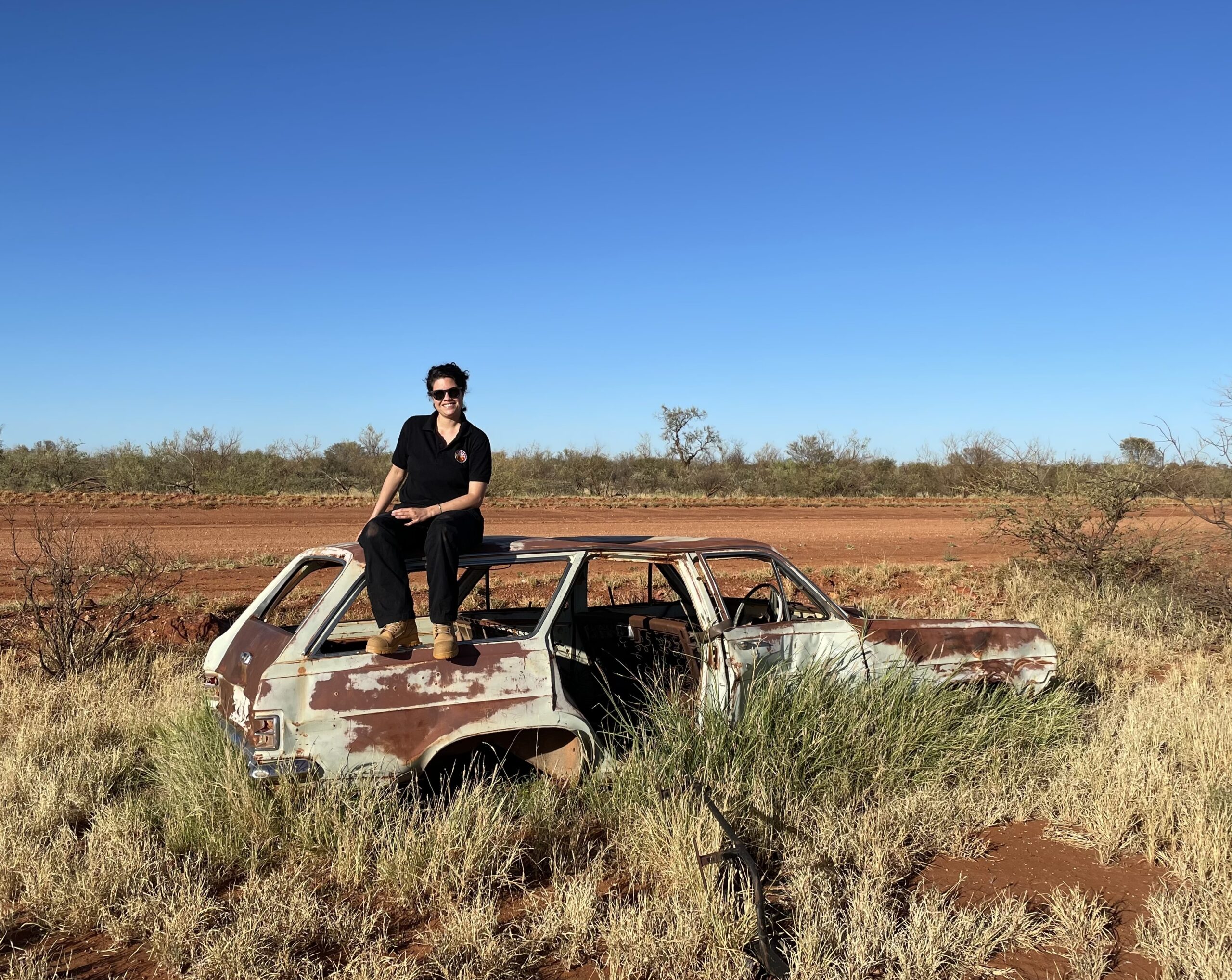 Woman sitting on top of an old rusted out car that has been abandoned on the side of a dirt road.