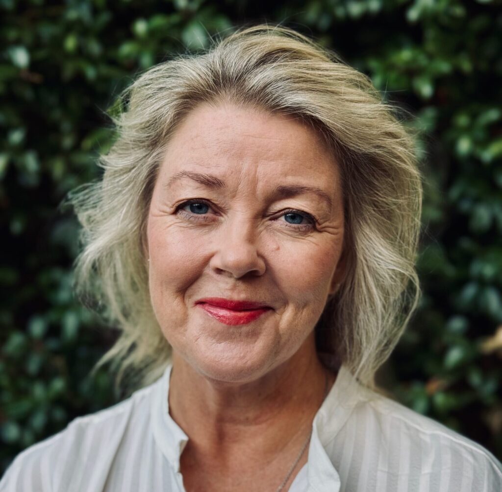 Woman with light hair in white shirt stands in front of lush green foliage background.
