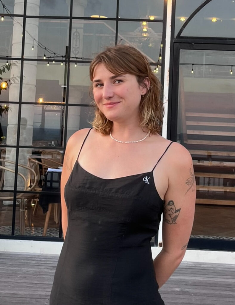 A woman in black CK top and pearl necklace stands in front of building with large windows and string lights.