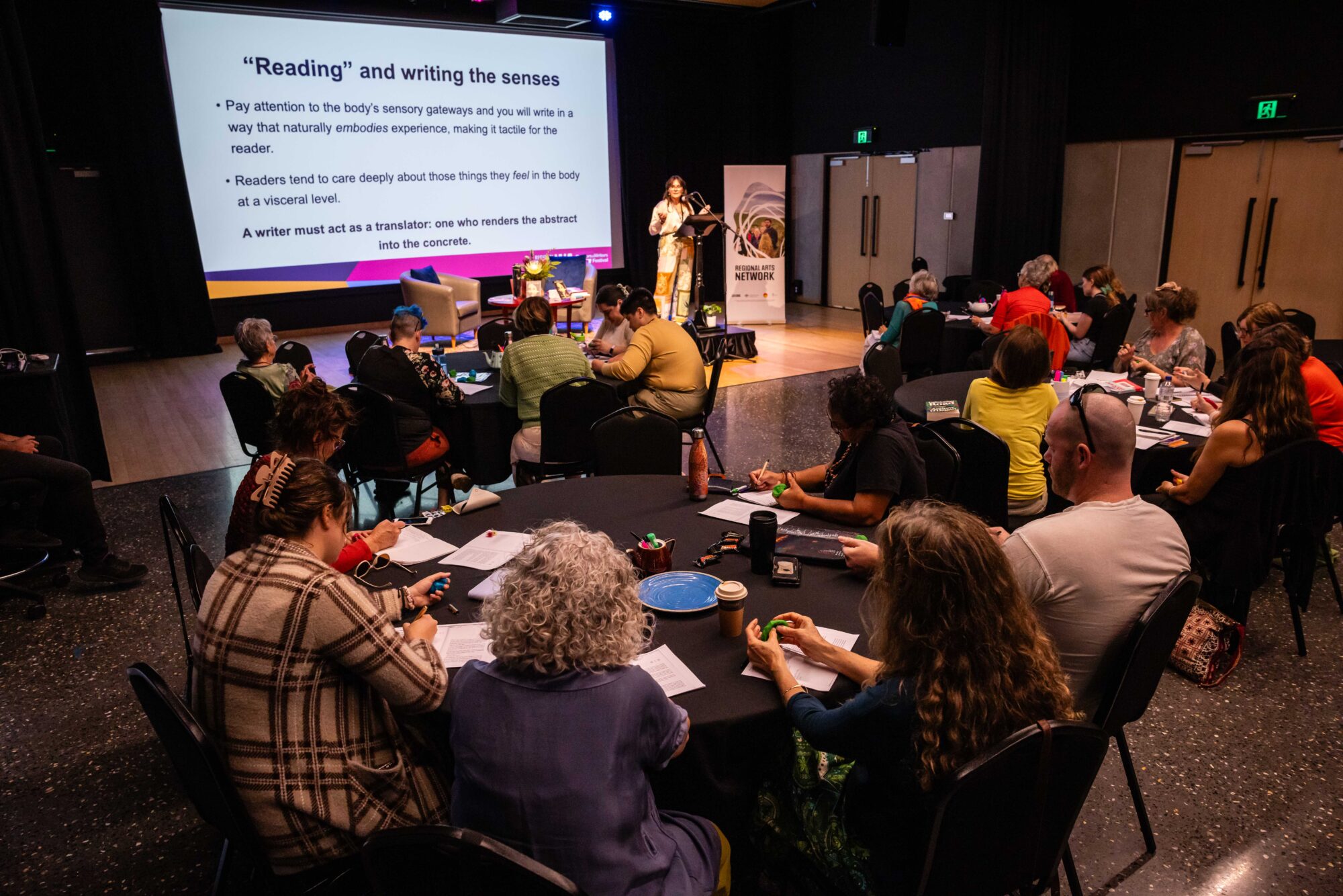 Audience seated at 4 round tables watching a presentation on writing and sensory signals; screen displays key points.