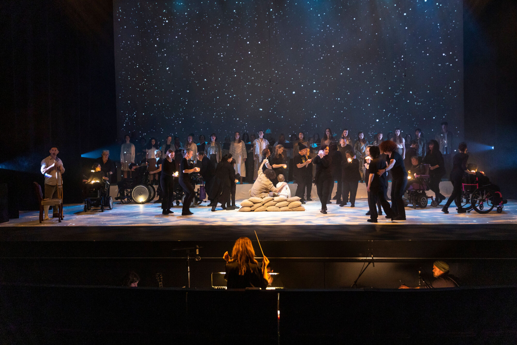 Stage performance under starry sky with black-clad performers and musicians; central figures lie on sandbags.