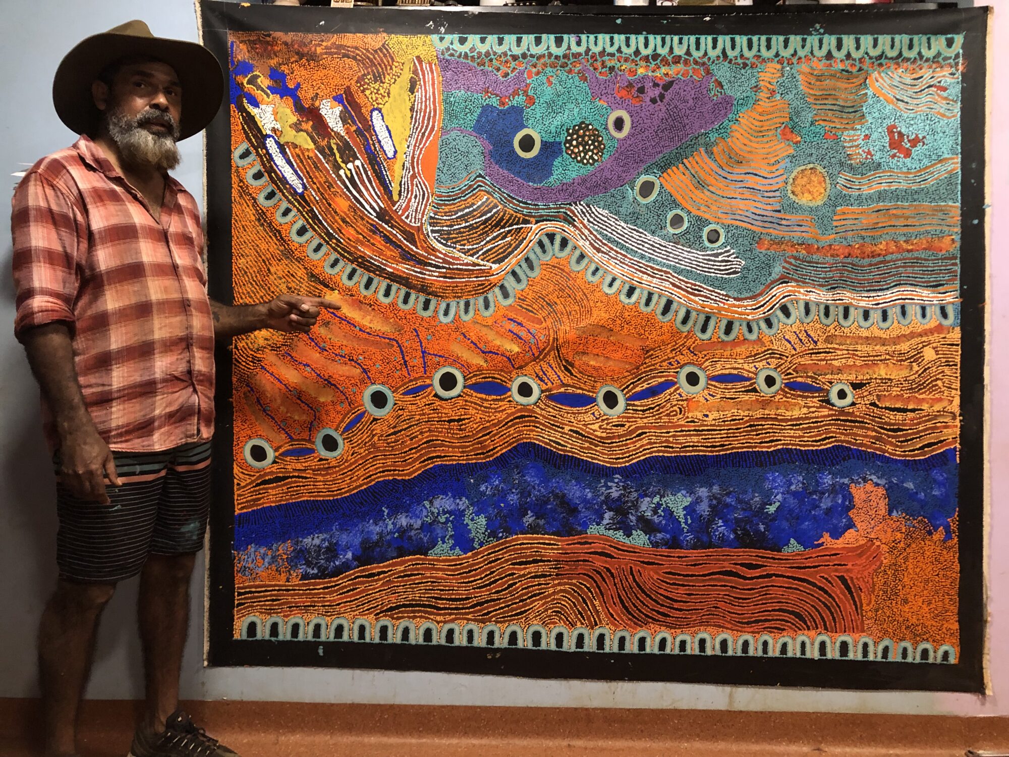 First Nations man wearing a hat standing next to and pointing at a large scale painting, inside