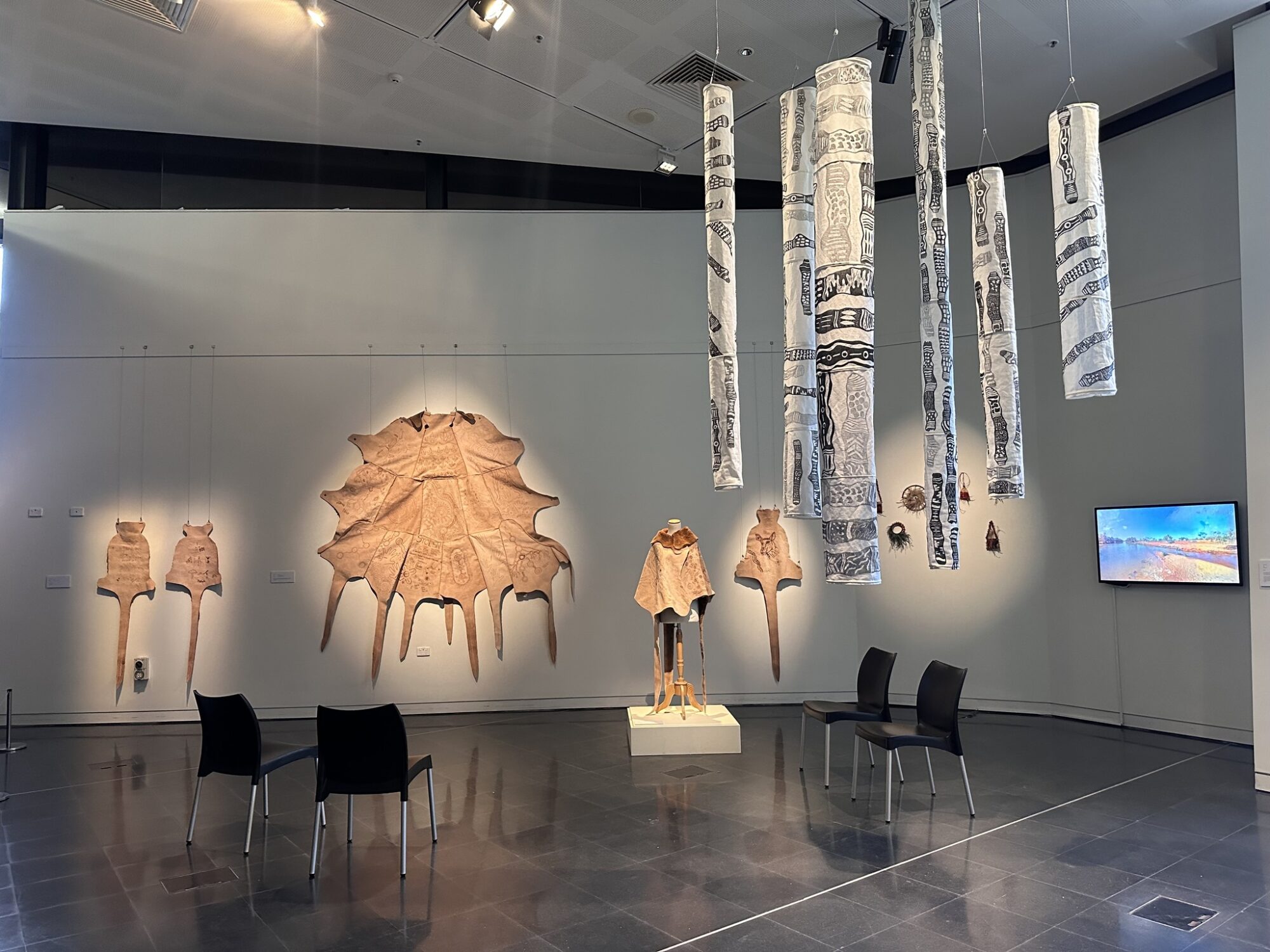 Gallery with textile art on walls and ceiling; mannequin on pedestal, chairs, and screen showing image or video.