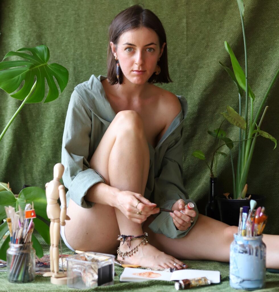 Artist Jenna Pickering wearing a green shirt, sitting in front of a green backdrop with plants and painting supplies