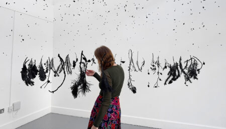 Katie Breckon, in green top and floral skirt stands before art installation of black suspended objects against white wall.