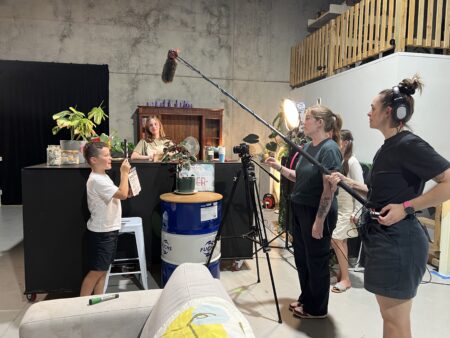 Behind-the-scenes of film shoot in indoor studio; crew with camera, boom mic, clapperboard, lights, and props.