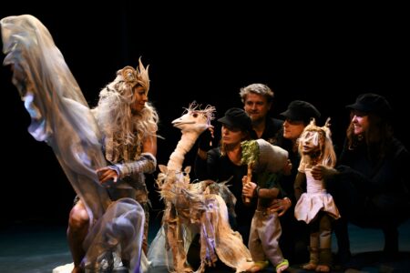 four people dressed in all black handle puppets in a dark setting on stage, one person wears a flowing costume