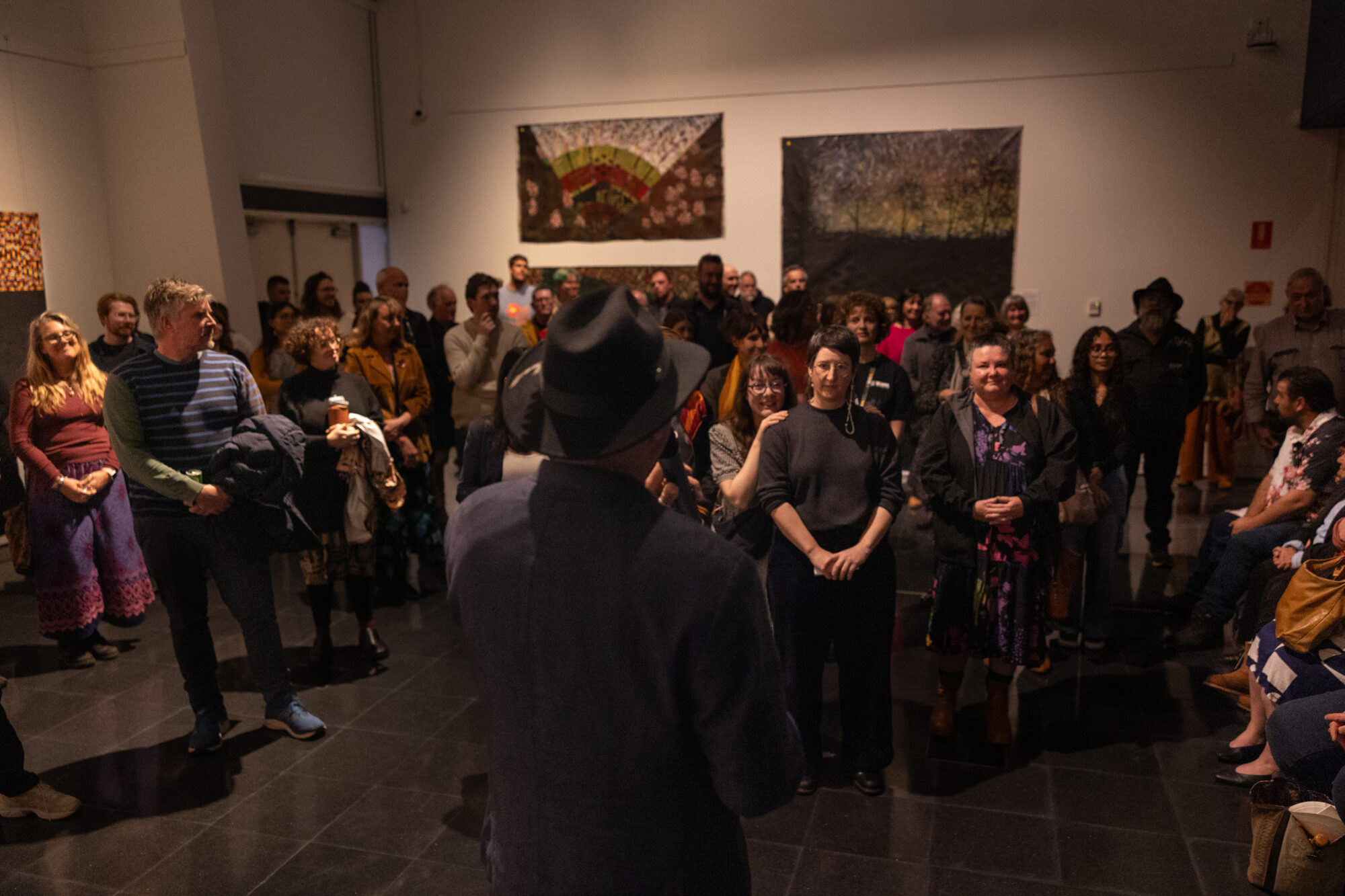 A man in a hat holding a microphone has his back turned to the camera, facing a full crowd of people, two large paintings hang on the wall behind the crowd.