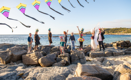 People dressed up in colourful costumes, standing on breakwater rocks, under a string of kites.