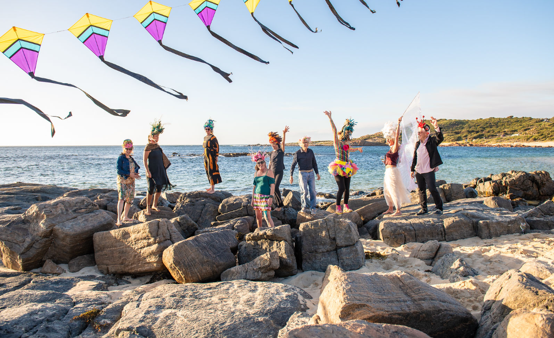 People dressed up in colourful costumes, standing on breakwater rocks, under a string of kites.