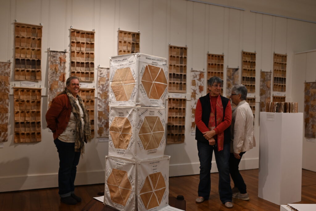 Three people walking through and engaging with an art exhibition. Textiles in various earthy-brown colours are displayed on the wall and plinths.