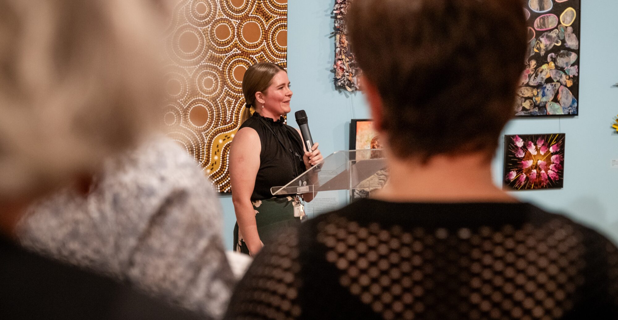 Woman talking into a microphone behind a podium in front of a crowd at an art gallery exhibition