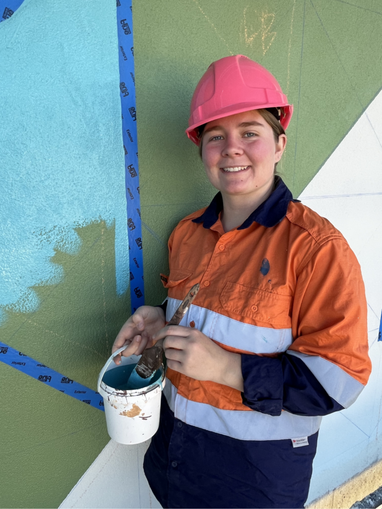 Woman wearing hi-vis and a hard hat, posing with a paint brush and paint bucket in front of a wall mural in progress.