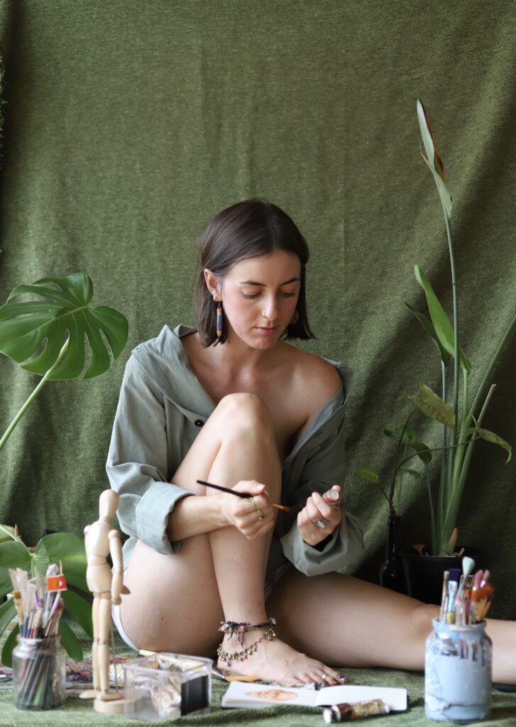 Woman sitting in front of a sage green backdrop surrounded but potted plants and panting supplies.