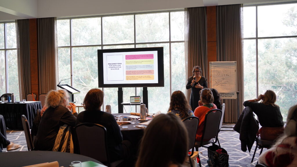 Woman presenting in front of a conference room of people seated in a banquet arrangement.