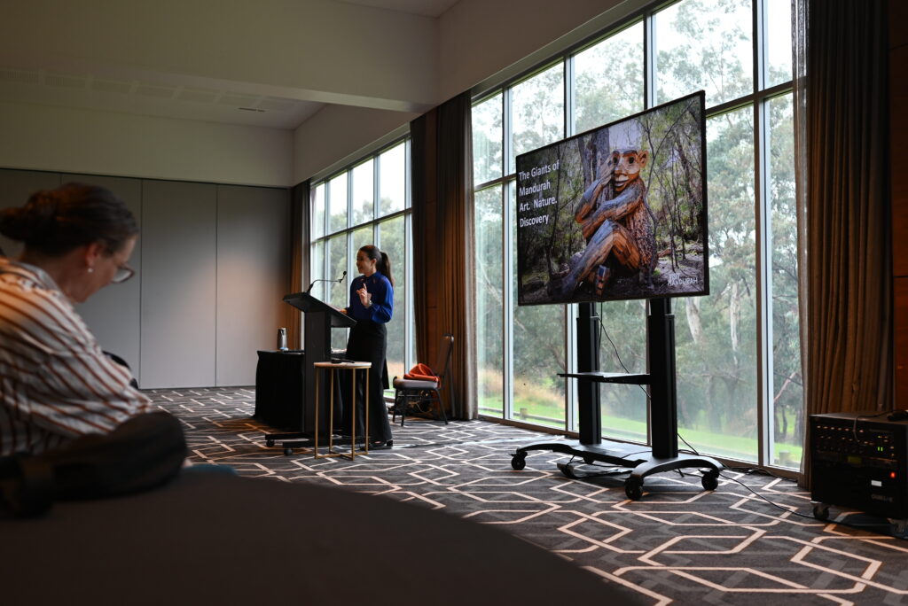 Woman presenting in front of a conference room of people seated in a banquet arrangement.