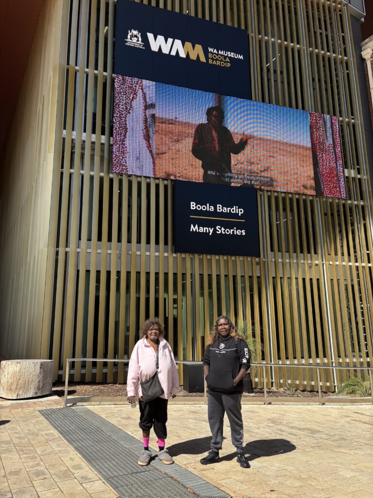 Two women standing in front of a museum with a large bilboard screen behind them.