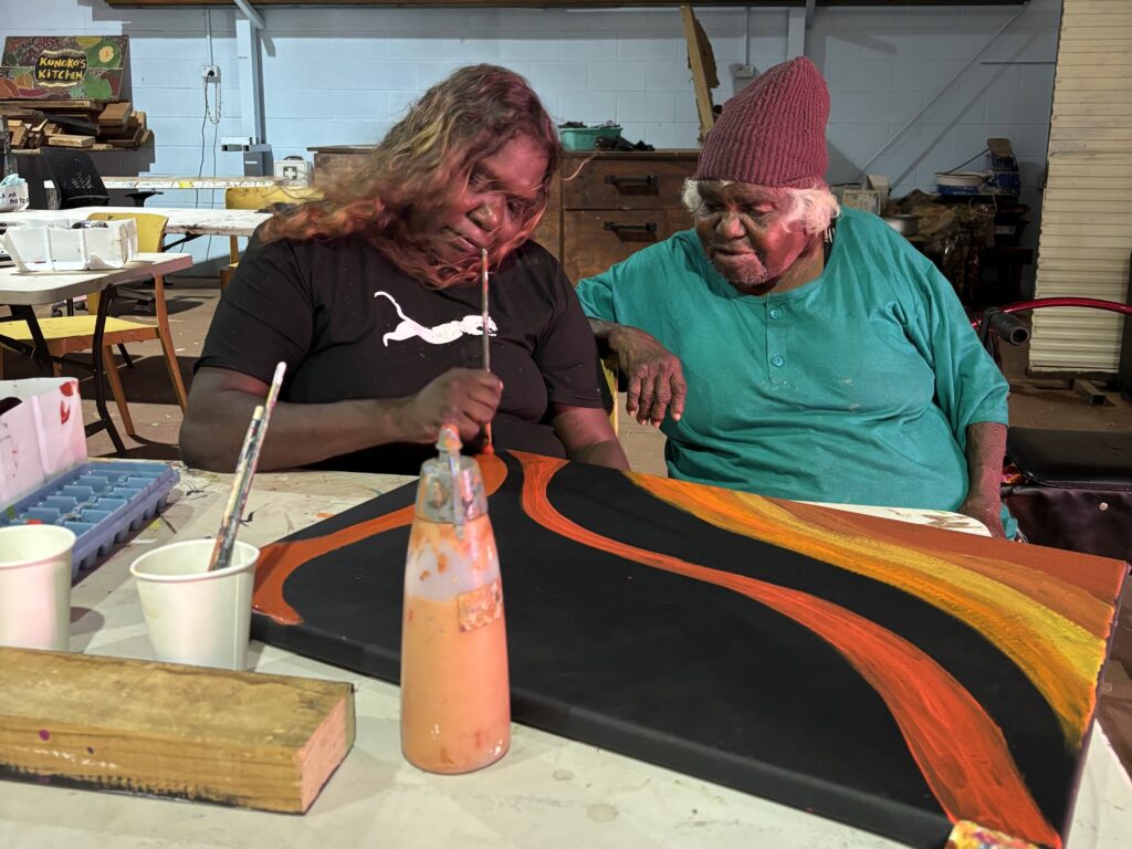 Two women sitting at a table in front of a painting in-progress. One of the women is actively painting on the canvas.